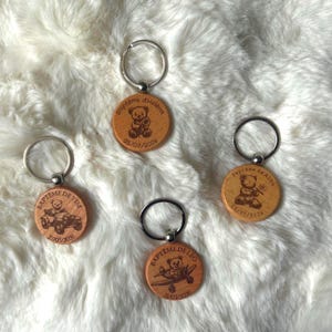 May include: Four circular wooden keychains with silver metal rings. Each keychain features a laser-engraved teddy bear illustration, a name, the word Bapteme, and a date. The designs include a bear with a flower, a bear in a car, and a bear in a plane.