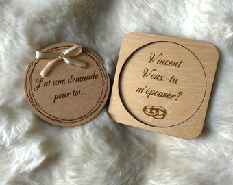 Puede incluir: Dos placas de madera clara con texto grabado. La placa circular tiene un lazo de cinta color crema y el texto J'ai une demande pour toi. La placa cuadrada presenta el texto Vincent Veux-tu m'épouser? junto a dos anillos.