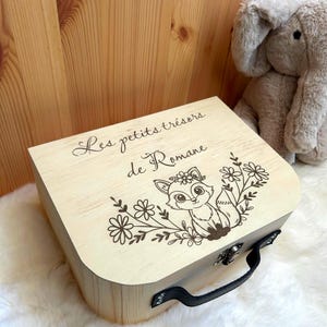 May include: A light-colored wooden box with a black handle and a metal latch. The lid features a laser-engraved illustration of a fox surrounded by flowers, along with the text "Les petits trésors de Romane" written in a cursive script font.