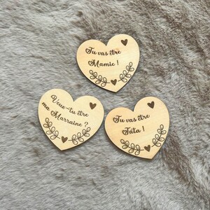 May include: Three light-colored wooden heart-shaped ornaments featuring laser-engraved text and botanical motifs. The text reads: Tu vas être Mamie!, Veux-tu être ma Marraine?, and Tu vas être Tata!, each decorated with small hearts and leaf sprigs.
