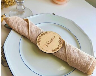 Puede incluir: Una servilleta de tela beige doblada sobre un plato blanco con borde azul. Un marcador de sitio redondo de madera se coloca sobre la servilleta, con un diseño de corona de laurel grabado a láser y el nombre Valentin en cursiva.