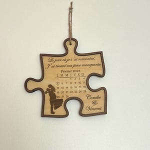 May include: A light-colored wooden puzzle piece wall hanging with dark brown edges, featuring a February 2018 calendar, a silhouette of a couple, the names Coralie and Vincent, and French text that reads: Le jour où je t'ai rencontré, J'ai trouvé ma pièce manquante.