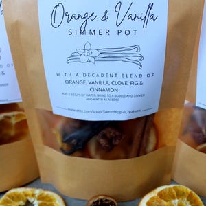May include: Three brown paper bags of simmer pot blend with the text "Orange & Vanilla". The bags contain dried orange slices, cinnamon sticks, cloves, and vanilla beans. The text on the label reads "With a decadent blend of orange, vanilla, clove, fig & cinnamon."