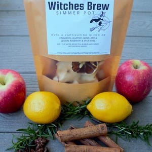 Könnte beinhalten: Ein brauner Papiersack mit der Aufschrift "Witches Brew Simmer Pot" mit einem transparenten Fenster, umgeben von roten Äpfeln, gelben Zitronen, Zimtstangen, Sternanis und Rosmarin. Der Beutel enthält eine Mischung aus Zimt, Piment, Nelken, Apfel, Zitrone, Rosmarin und Sternanis.