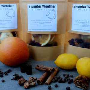 Könnte beinhalten: Drei braune Papiertüten mit Klarsichtfenstern, beschriftet mit "Sweater Weather Simmer Pot". Die Tüten enthalten Trockenfrüchte und Gewürze. Eine große orangefarbene Grapefruit und zwei gelbe Zitronen liegen davor, zusammen mit Zimtstangen und Pfefferkörnern.