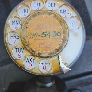 Puede incluir: Un dial de teléfono giratorio vintage con una cara circular blanca, letras negras y números rojos. El centro muestra una etiqueta de color canela con el número escrito a mano 898-5430 y el logotipo de GTE. El dial está cubierto por una tapa de plástico transparente.