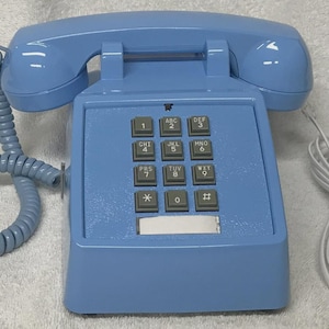 May include: A light blue landline telephone with a matching handset and a coiled cord. The base features a standard twelve-button keypad with grey square buttons, each marked with numbers and letters, and a blank rectangular label area at the bottom.