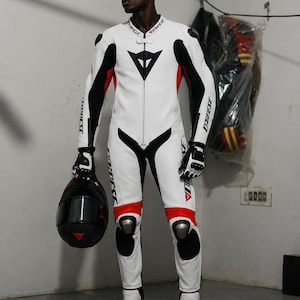 May include: White and black motorcycle racing suit with red accents. The suit features a black triangle logo on the chest and knee pads. The person is holding a black helmet with a red triangle logo.