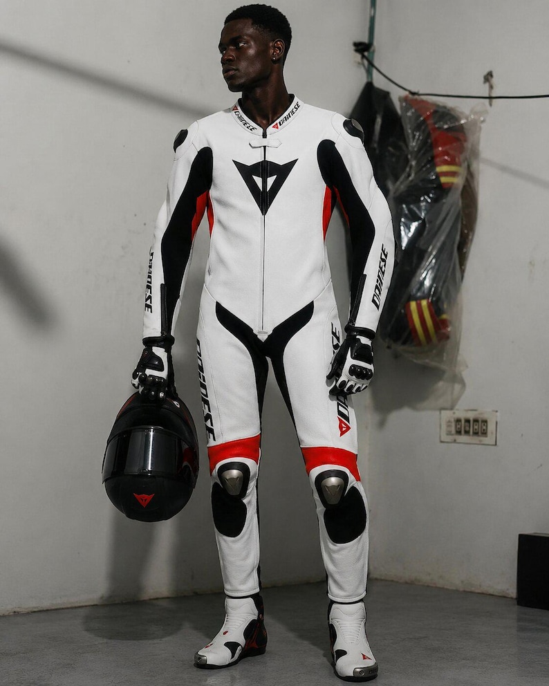 May include: White and black motorcycle racing suit with red accents. The suit features a black triangle logo on the chest and knee pads. The person is holding a black helmet with a red triangle logo.