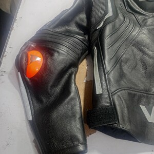 Puede incluir: Chaqueta de moto de cuero negro con detalles plateados y un protector de codo naranja. La chaqueta presenta un dise&ntilde;o acolchado en hombros y brazos, con cierre de velcro en la mu&ntilde;eca. Se aprecia el logotipo V.