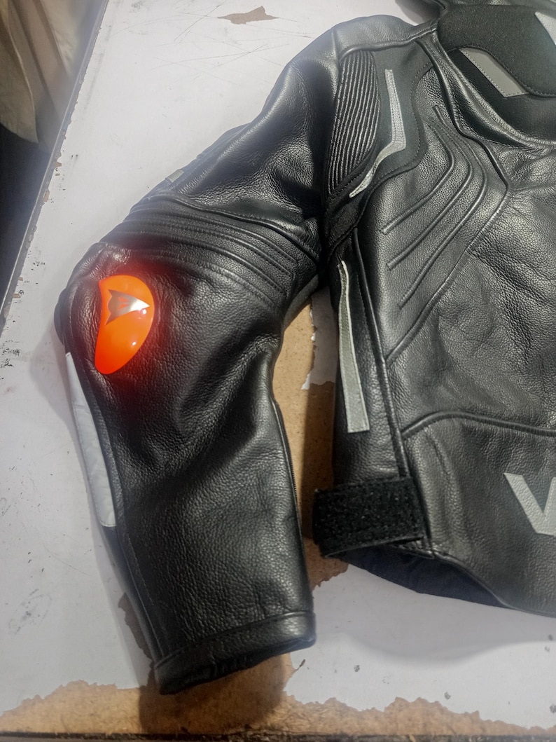 Puede incluir: Chaqueta de moto de cuero negro con detalles plateados y un protector de codo naranja. La chaqueta presenta un dise&ntilde;o acolchado en hombros y brazos, con cierre de velcro en la mu&ntilde;eca. Se aprecia el logotipo V.