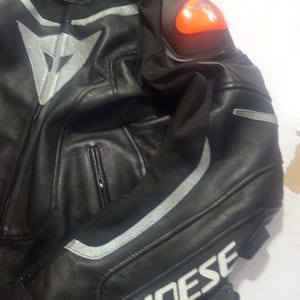 Puede incluir: Chaqueta de moto de cuero negro con detalles plateados y el nombre de la marca "DAINESE" en blanco. Incluye un protector de hombro naranja y cierre de cremallera. La chaqueta est&aacute; dise&ntilde;ada para protecci&oacute;n y estilo.