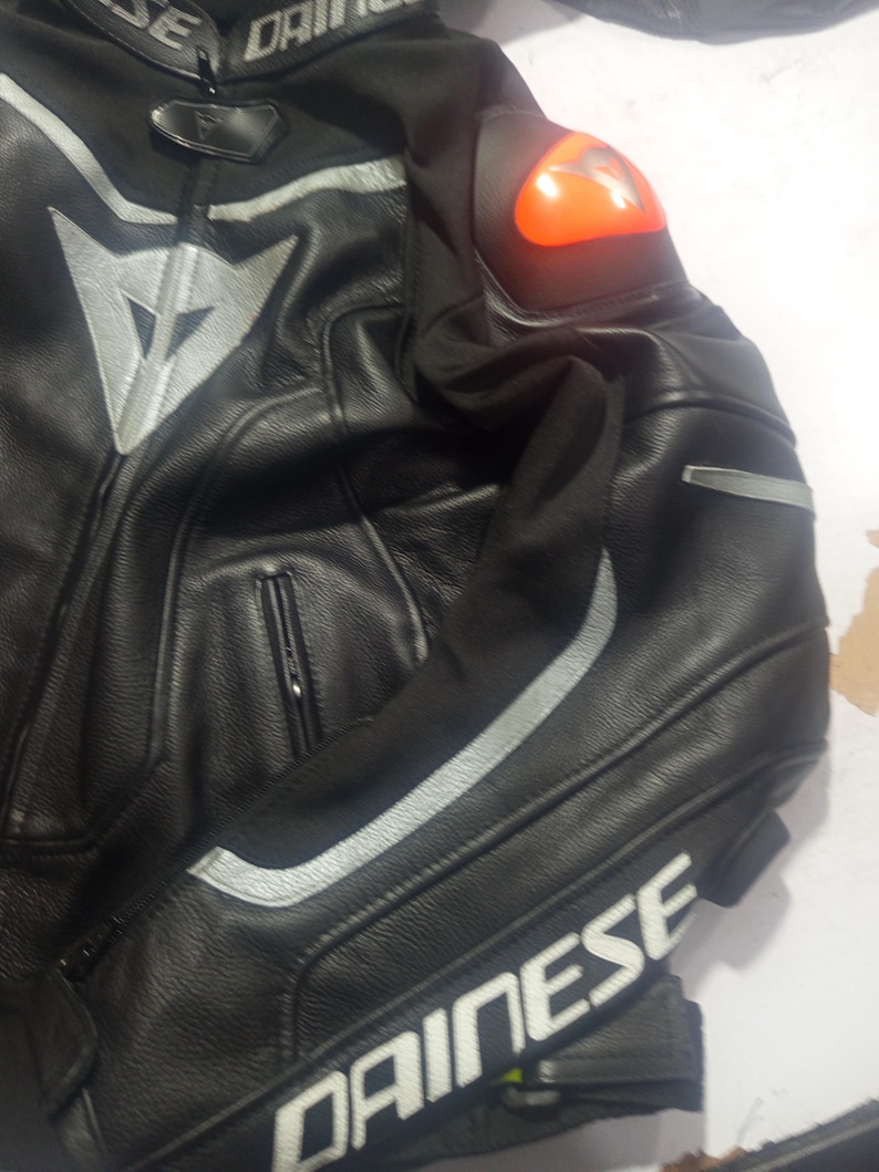 Puede incluir: Chaqueta de moto de cuero negro con detalles plateados y el nombre de la marca "DAINESE" en blanco. Incluye un protector de hombro naranja y cierre de cremallera. La chaqueta est&aacute; dise&ntilde;ada para protecci&oacute;n y estilo.