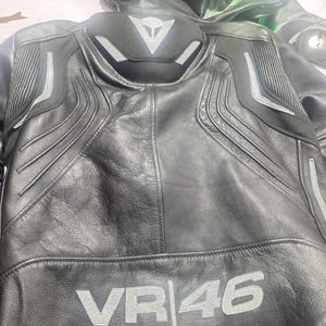 Puede incluir: Chaqueta de moto de cuero negro con detalles plateados y el texto "VR|46" en plata. La chaqueta tiene un cuello alto y hombros acolchados. Otra chaqueta de moto es visible en el fondo.