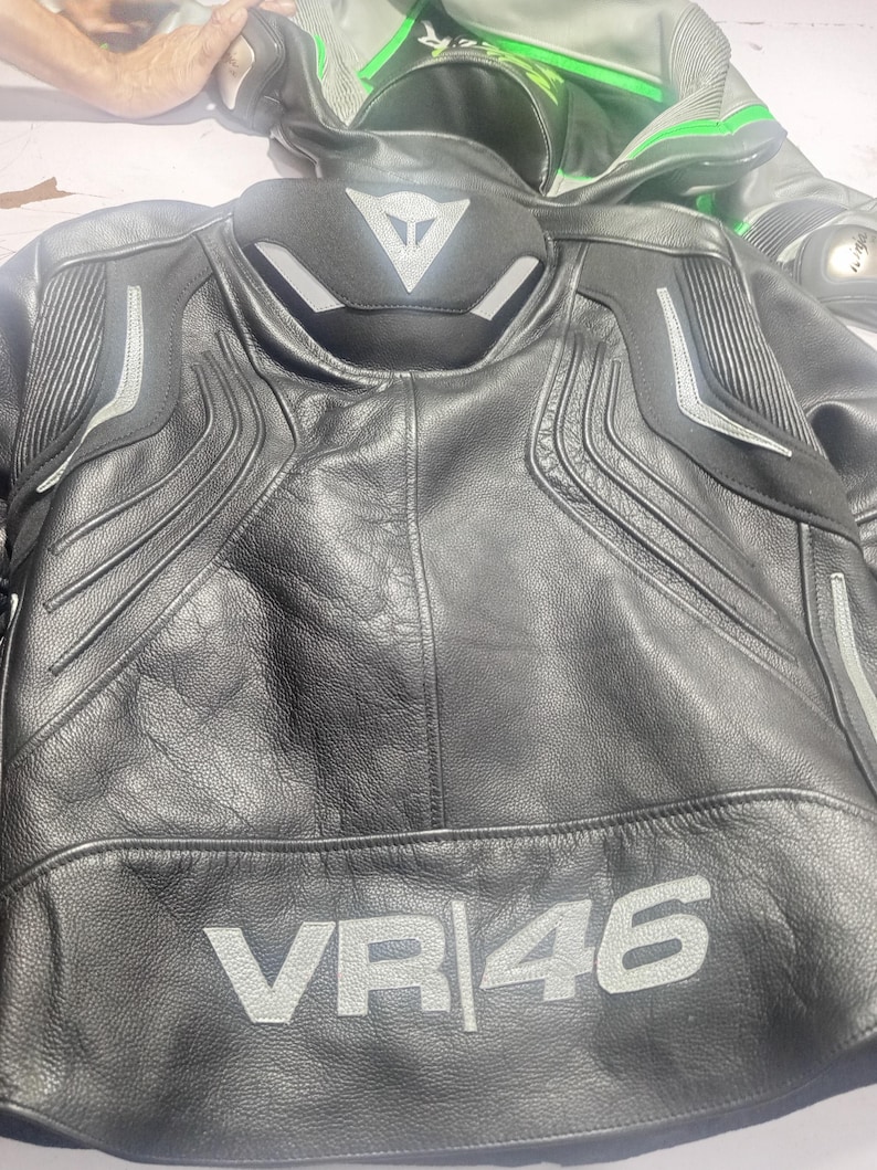 Puede incluir: Chaqueta de moto de cuero negro con detalles plateados y el texto "VR|46" en plata. La chaqueta tiene un cuello alto y hombros acolchados. Otra chaqueta de moto es visible en el fondo.