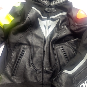 Puede incluir: Chaqueta de moto de cuero negro con detalles plateados y el logotipo de Dainese. La chaqueta tiene cremallera frontal, bolsillos y hombros acolchados. En el fondo se ven otras chaquetas, una con detalles verdes y blancos y otra con detalles amarillos.
