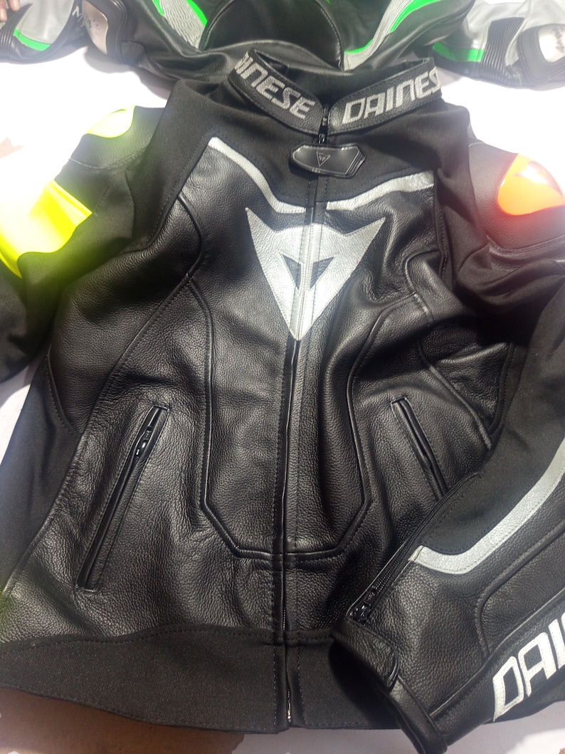 Puede incluir: Chaqueta de moto de cuero negro con detalles plateados y el logotipo de Dainese. La chaqueta tiene cremallera frontal, bolsillos y hombros acolchados. En el fondo se ven otras chaquetas, una con detalles verdes y blancos y otra con detalles amarillos.