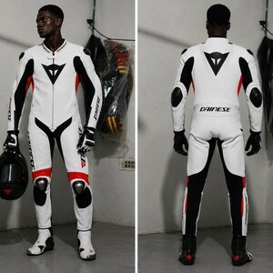 May include: White and black motorcycle racing suit with red accents. The suit features the Dainese logo and is worn with black gloves and boots. A black helmet is held in one hand. The suit has protective padding on the knees and elbows.