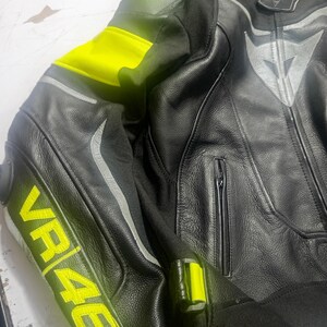 Puede incluir: Chaqueta de moto de cuero negro con detalles en amarillo ne&oacute;n en hombros y mangas. La chaqueta presenta el texto "VR46" en amarillo en la manga. Tiene cierre de cremallera y rayas reflectantes plateadas.