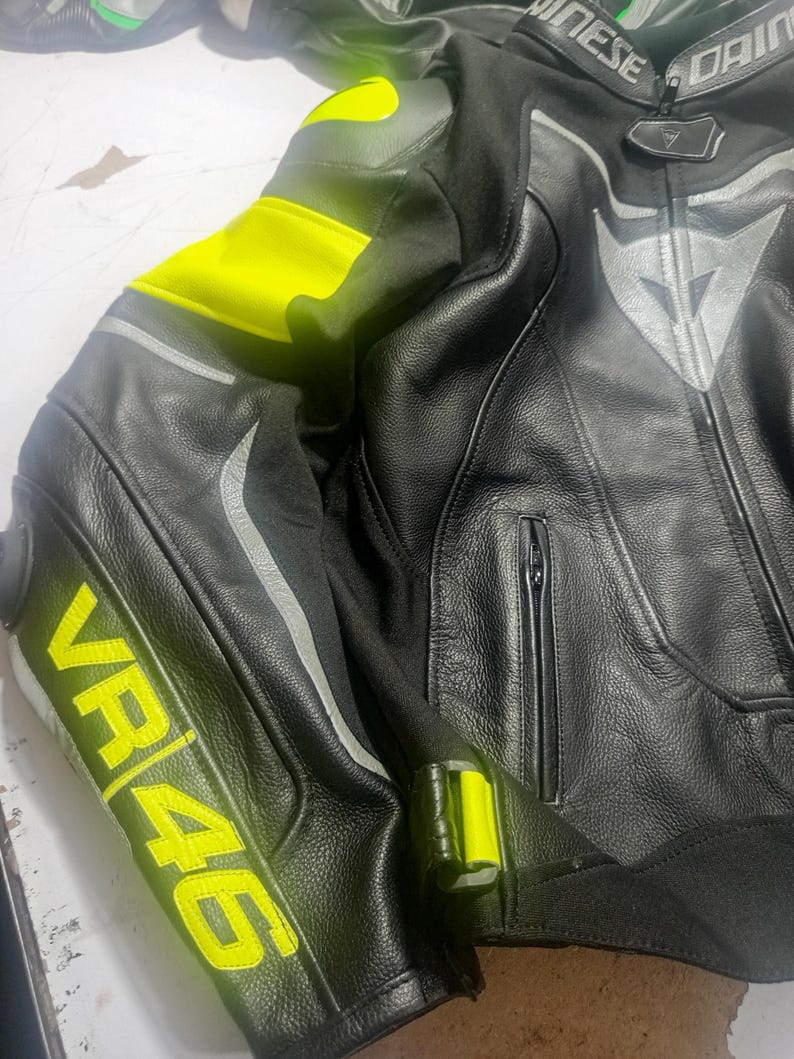 Puede incluir: Chaqueta de moto de cuero negro con detalles en amarillo ne&oacute;n en hombros y mangas. La chaqueta presenta el texto "VR46" en amarillo en la manga. Tiene cierre de cremallera y rayas reflectantes plateadas.