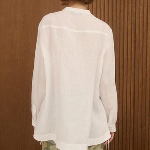 May include: A white long-sleeve shirt with a mandarin collar. The shirt has a relaxed fit and buttoned cuffs. The model is wearing patterned pants with a green and beige design. The background is a wooden wall.