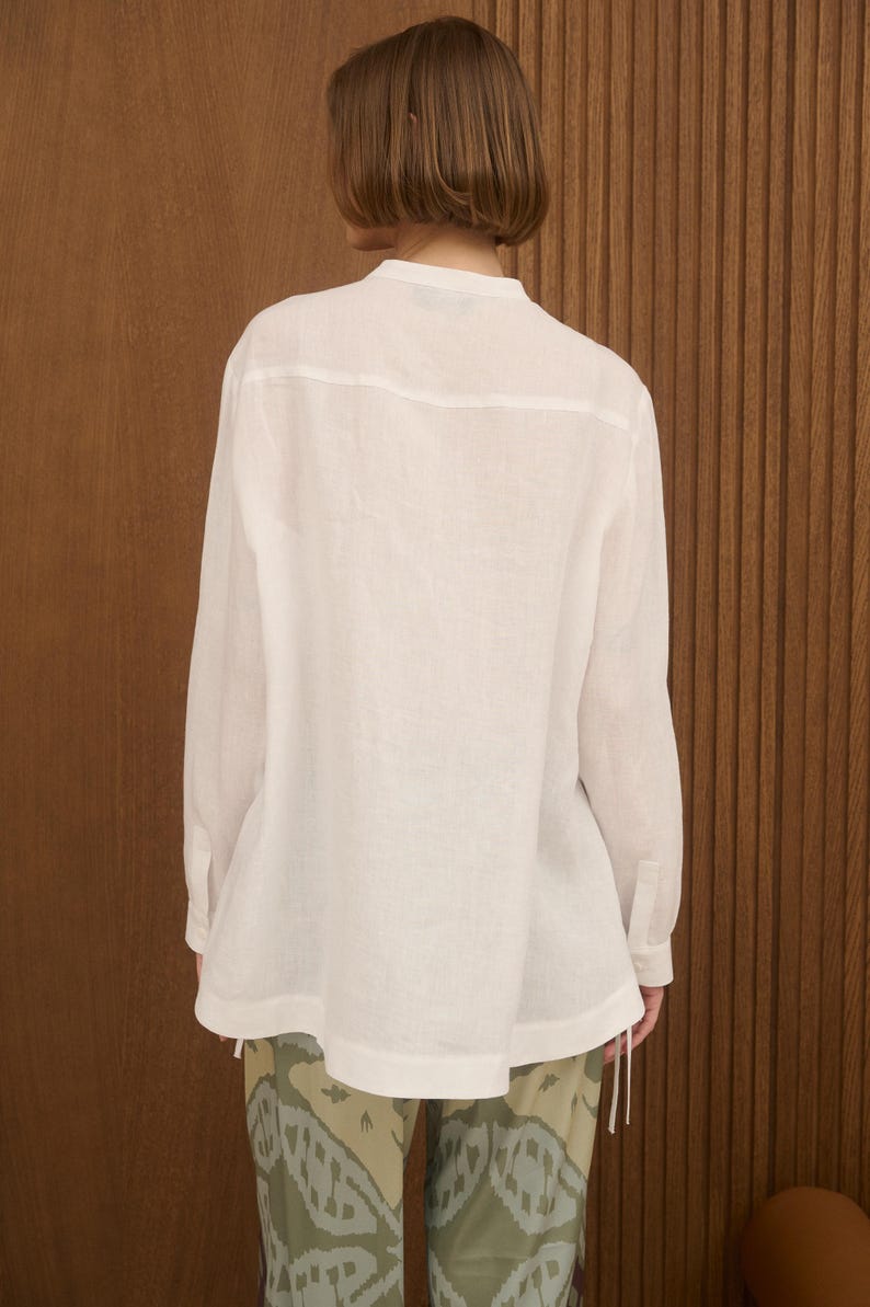 May include: A white long-sleeve shirt with a mandarin collar. The shirt has a relaxed fit and buttoned cuffs. The model is wearing patterned pants with a green and beige design. The background is a wooden wall.