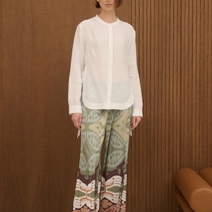 May include: A woman wearing a white long-sleeve shirt and patterned pants. The pants feature a design in shades of green, brown, and beige. The shirt has a mandarin collar and a button-down front. The outfit is paired with brown sandals.