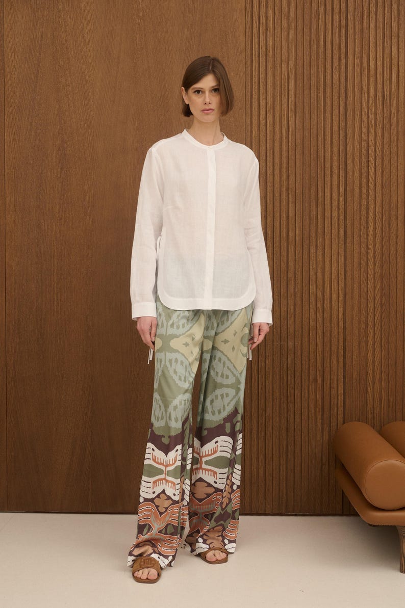 May include: A woman wearing a white long-sleeve shirt and patterned pants. The pants feature a design in shades of green, brown, and beige. The shirt has a mandarin collar and a button-down front. The outfit is paired with brown sandals.