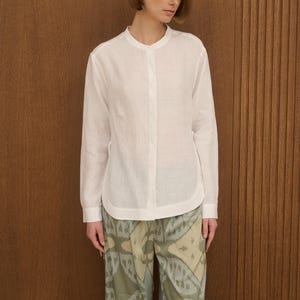 May include: A white long-sleeve shirt with a mandarin collar and button-down front. The shirt is paired with patterned pants in shades of green, brown, and beige. The outfit is simple and elegant.