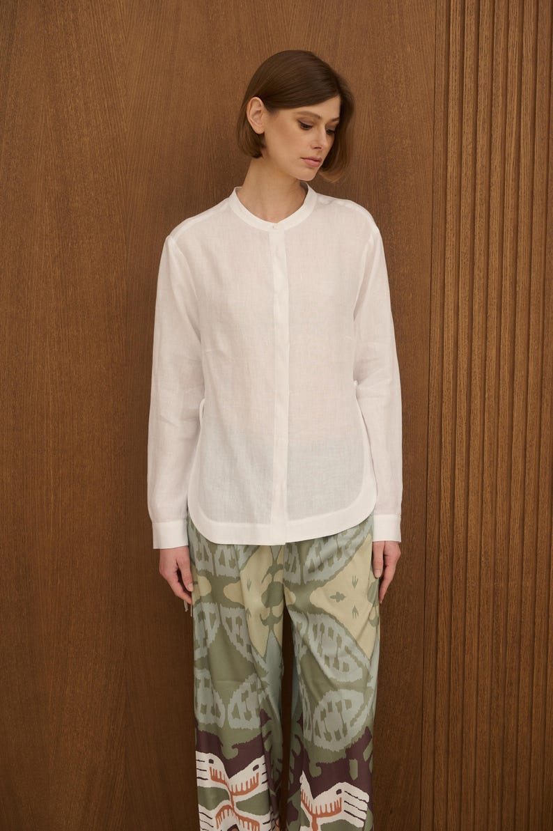 May include: A white long-sleeve shirt with a mandarin collar and button-down front. The shirt is paired with patterned pants in shades of green, brown, and beige. The outfit is simple and elegant.