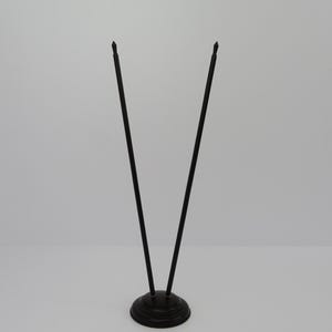 May include: A black metal table number holder with two slender, pointed rods extending upwards from a circular base. The base has a decorative, ridged design. The rods are angled slightly away from each other.
