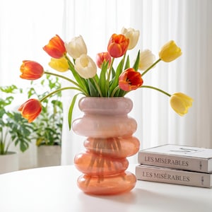 May include: A decorative glass vase with a stacked, rounded design, filled with colorful tulips. The tulips are a mix of yellow, orange, and white, creating a vibrant floral arrangement. The vase sits on a white table.