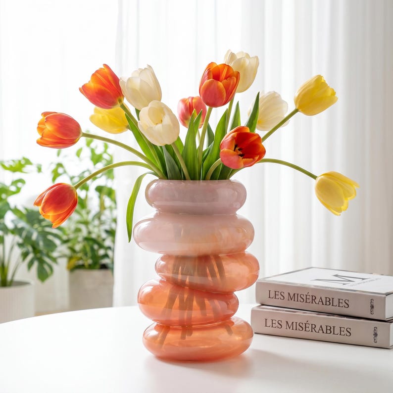 May include: A decorative glass vase with a stacked, rounded design, filled with colorful tulips. The tulips are a mix of yellow, orange, and white, creating a vibrant floral arrangement. The vase sits on a white table.