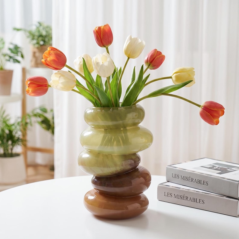 May include: A decorative vase with a stacked, rounded design in shades of green, brown, and orange. The vase holds a bouquet of tulips in orange, yellow, and white. The vase is on a white table, with a book titled "Les Miserables" in the background.
