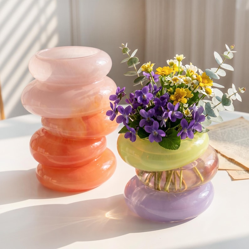 May include: Two decorative glass vases. One is stacked with pink and orange rings. The other vase is stacked with green, pink, and purple rings, and filled with purple, yellow, and white flowers and greenery.
