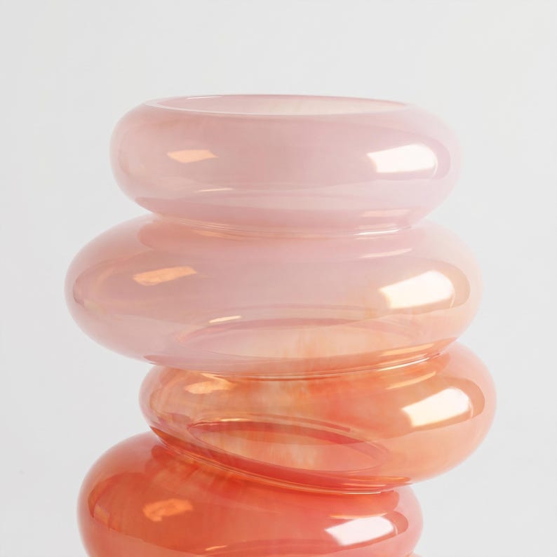 May include: A stacked glass vase in shades of pink and orange. The vase has a unique, rounded shape, with each section slightly smaller than the one below. The glass has a glossy finish, reflecting light and creating a soft, warm glow.