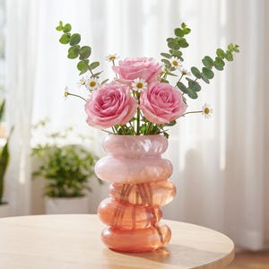 May include: A decorative vase with a stacked, rounded design in shades of pink and orange holds a bouquet of pink roses, small white daisies, and green eucalyptus leaves. The vase sits on a light-colored wooden table.