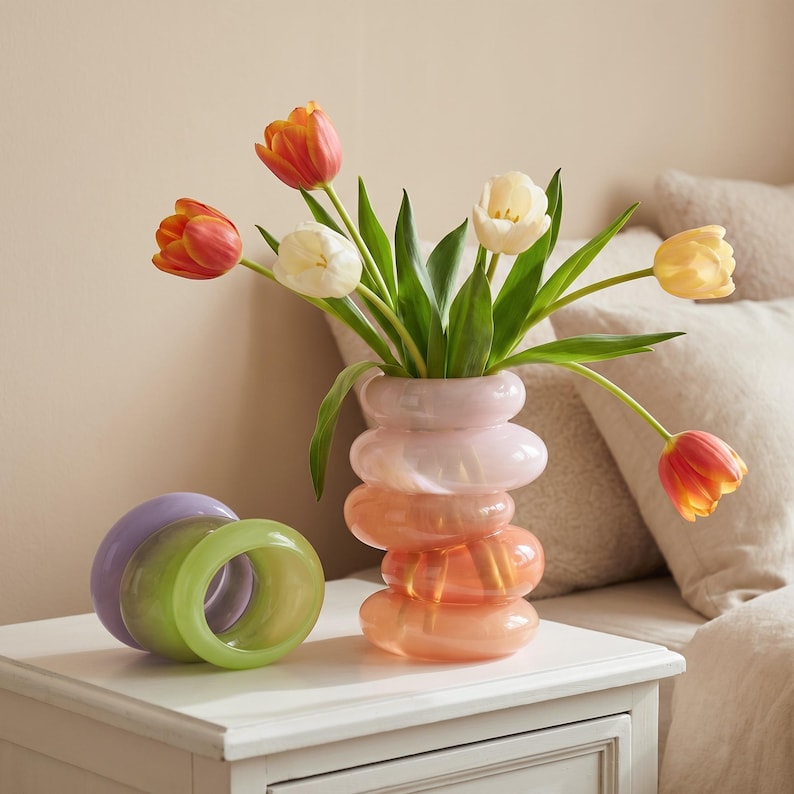 May include: A decorative vase with a stacked, ring-like design in shades of pink and orange holds a bouquet of tulips. Two decorative ring-shaped objects in green and purple sit on a white nightstand.