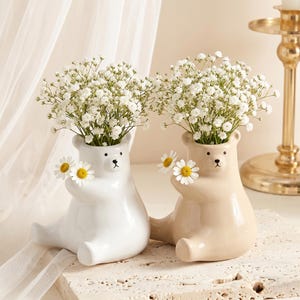 May include: Two ceramic bear-shaped vases, one white and one beige, each holding a bouquet of white flowers. The vases have black eyes and a small black nose. Daisies are held in the bear's arms. The vases are sitting on a stone surface.