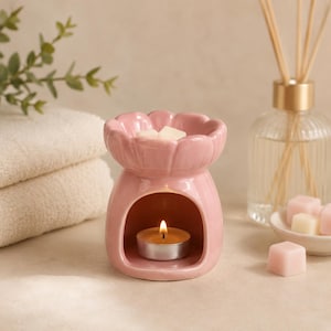 May include: A light pink ceramic wax warmer shaped like a flower with a scalloped rim. The base features a hollow archway containing a small lit tea light candle. A white wax melt sits in the top bowl, and small pink wax cubes are nearby.