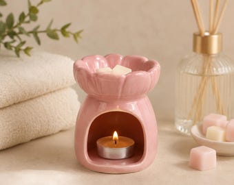 May include: A light pink ceramic wax warmer shaped like a flower with a scalloped rim. The base features a hollow archway containing a small lit tea light candle. A white wax melt sits in the top bowl, and small pink wax cubes are nearby.
