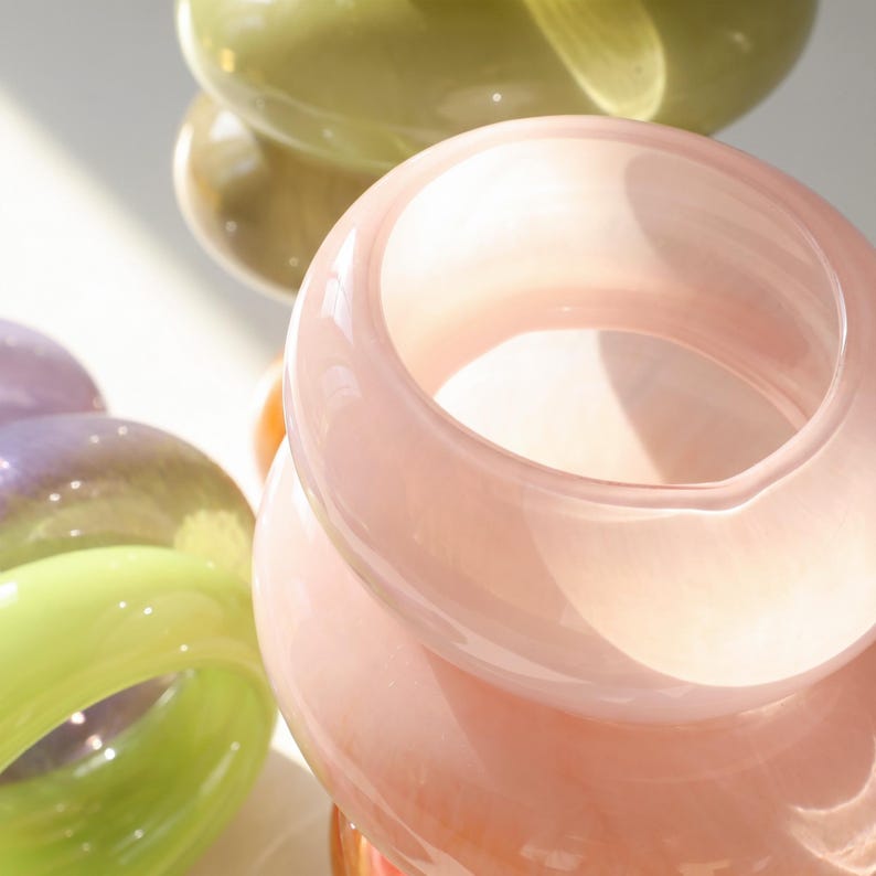 May include: Close-up of several colorful glass vases. The vases are in shades of pink, green, and purple. The glass is smooth and glossy, with a rounded shape. The light reflects off the glass, creating highlights and shadows.