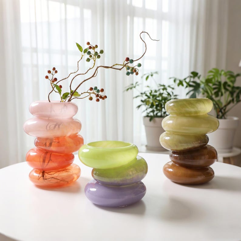 May include: Three stacked glass vases in shades of pink, orange, green, purple, and brown. The vases have a unique, rounded design. One vase holds a decorative branch with berries. The vases are on a white table.