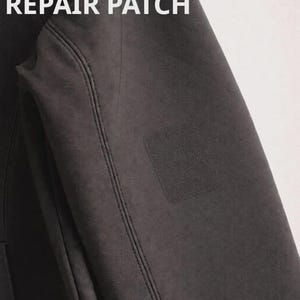 Peut inclure: Gros plan sur une manche de veste gris foncé avec un patch de réparation carré. Le texte "REPAIR PATCH" est affiché en lettres blanches en haut de la manche. La veste a des coutures visibles.