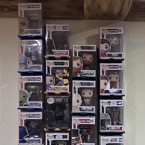 Peut inclure: Une collection de figurines Funko Pop! exposée sur un mur. Les figurines sont dans leurs boîtes d'origine, présentant divers personnages d'une franchise populaire. Les boîtes transparentes permettent de voir les figurines. L'ensemble est fixé sur un mur de couleur neutre.