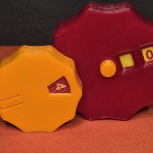 Peut inclure: Deux objets octogonaux imprimés en 3D. L'un est jaune avec un indicateur "4", l'autre est rouge avec un indicateur "0". Les objets ont un design en forme de roue dentée et sont probablement des composants d'un mécanisme ou d'un appareil plus grand.