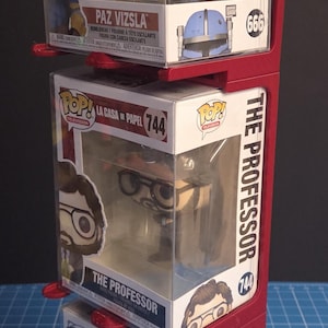 Peut inclure: Un présentoir rouge à trois niveaux contenant trois figurines Funko Pop! Les figurines sont dans leurs boîtes d'origine, avec des personnages de Star Wars et La Casa de Papel. Le présentoir est sur une surface quadrillée bleue.