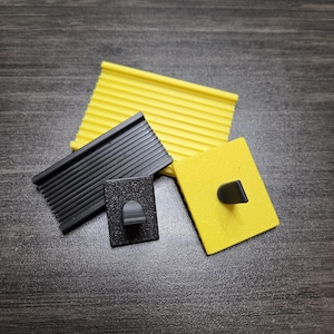 Puede incluir: Cuatro pequeños objetos de plástico texturizado en negro y amarillo están dispuestos sobre una superficie oscura con vetas de madera. Los objetos incluyen una pieza rectangular con crestas paralelas, un cuadrado con un asa curva y un cuadrado con un asa negra.