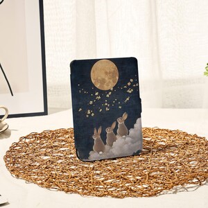 Può includere: Una custodia per tablet blu scuro con un'illustrazione stravagante di tre conigli che guardano una luna piena. Il design include nuvole bianche e accenti floreali dorati, creando una scena notturna serena.