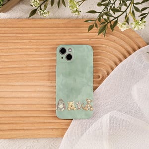 以下が含まれることがあります： ミントグリーンの携帯電話ケース。水彩画風のデザインで、イーヨー、プーさん、ピグレット、ティガーなどの漫画のキャラクターが下部に並んでいます。ケースにはカメラとボタン用の切り欠きがあります。